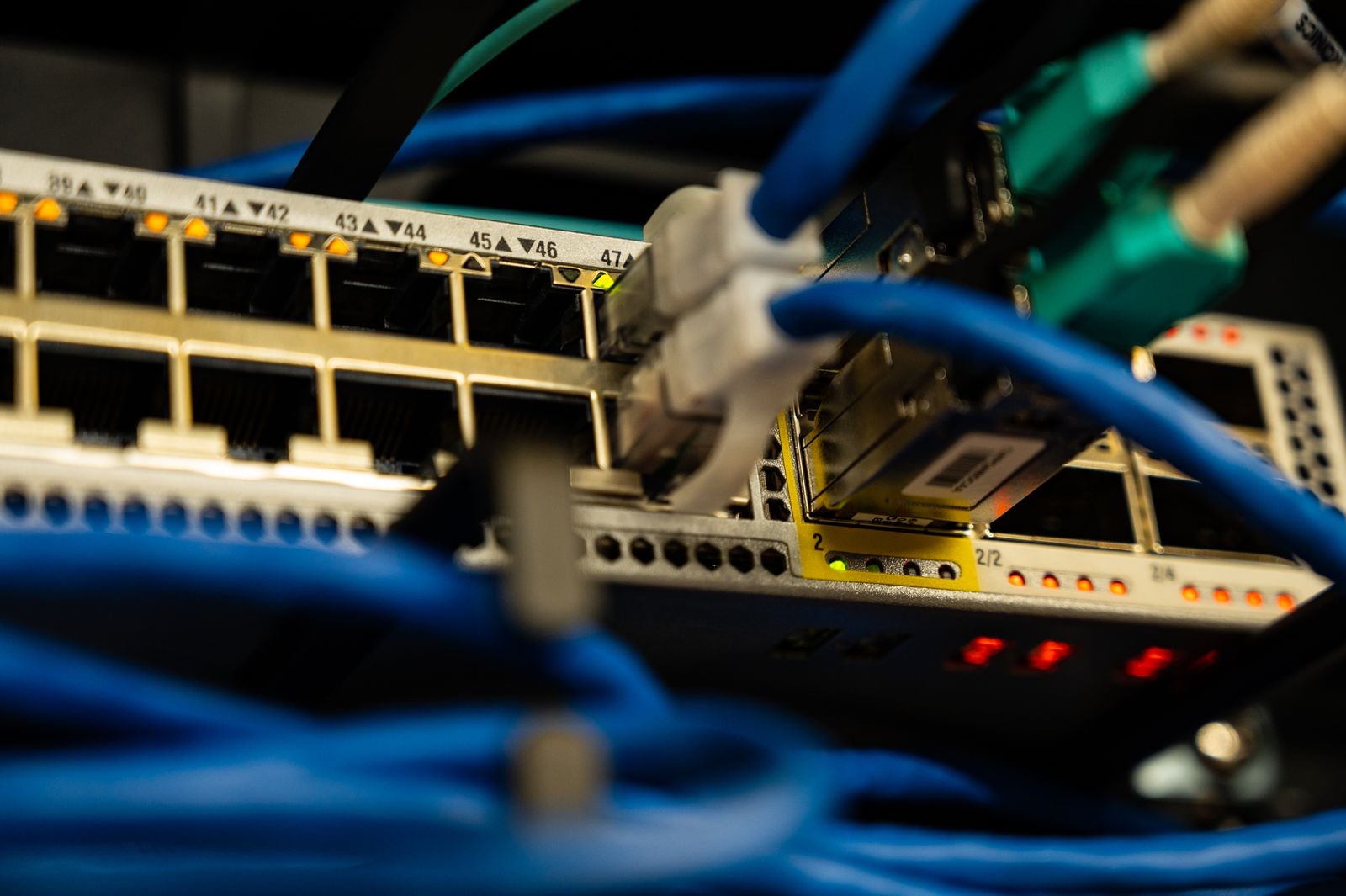 Cận cảnh mặt trước một switch mạng nhiều cổng RJ45 mạ vàng đánh số 39 đến 47, một hàng đèn báo cam đang sáng, các sợi cáp mạng xanh dương cuộn ở tiền cảnh cùng vài đầu nối xanh ngọc và đèn LED đỏ phía dưới, nền tối