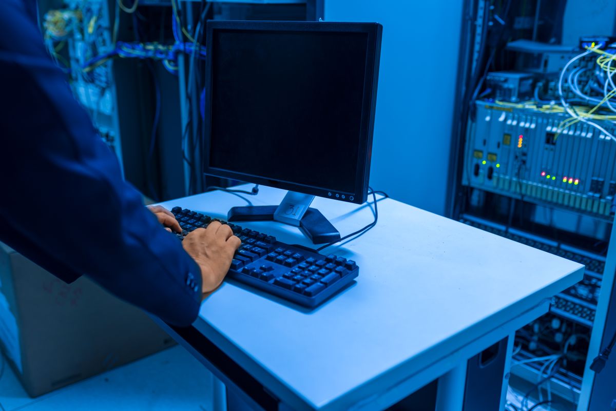 A technician standing and typing at a monitor on a white desk inside a blue-lit server room, with network cabinets and cable bundles behind